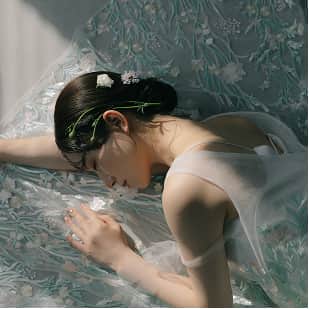 淡い光の中で、花飾りをつけた女性がベッドに横たわり、薄い衣装に包まれている。