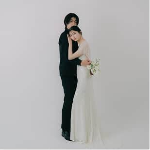 白い背景の前で、花束を持ったウェディングドレス姿の女性とタキシードの男性が親密に立っている。